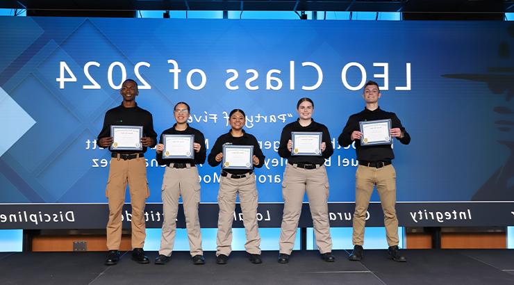 A group of 5 Law Enforcement Option students standing in a line in front of a large blue screen. 屏幕上用白色文字写着2024届LEO班. All students are holding their graduation certificate while wearing black, 长袖有领衬衫, 卡其色的裤子, 黑色的鞋子.